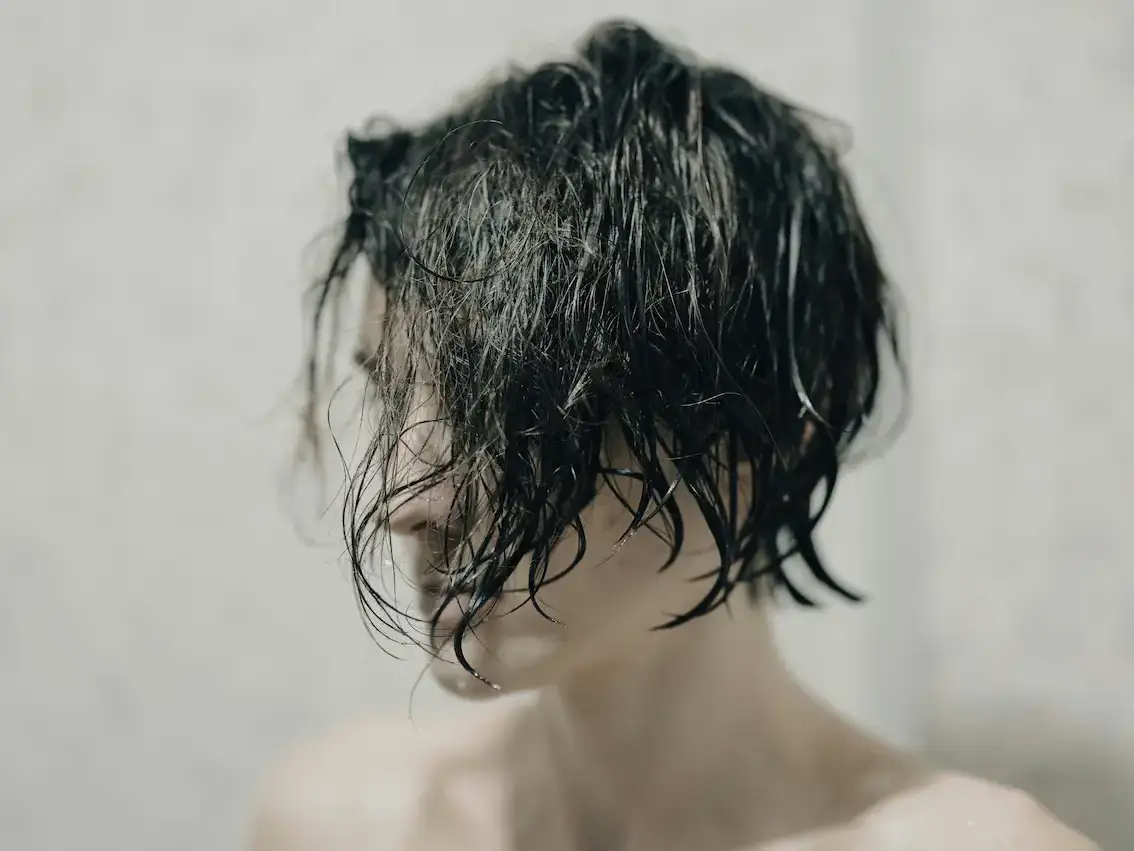 Woman with wet, dark hair falling across her face after washing, photographed against a neutral grey background.