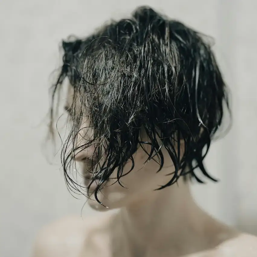 Woman with wet, dark hair falling across her face after washing, photographed against a neutral grey background.
