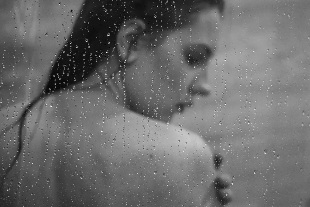 Woman seen through a glass shower screen covered in water droplets, with wet hair, in black and white.