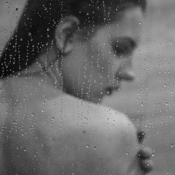 Woman seen through a glass shower screen covered in water droplets, with wet hair, in black and white.