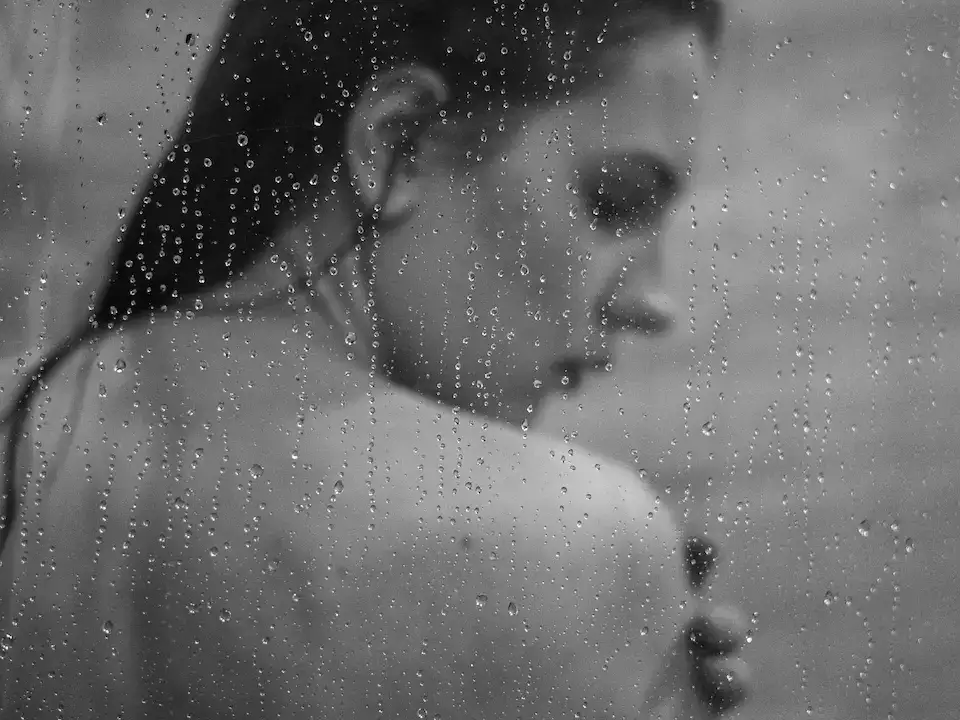 Woman seen through a glass shower screen covered in water droplets, with wet hair, in black and white.