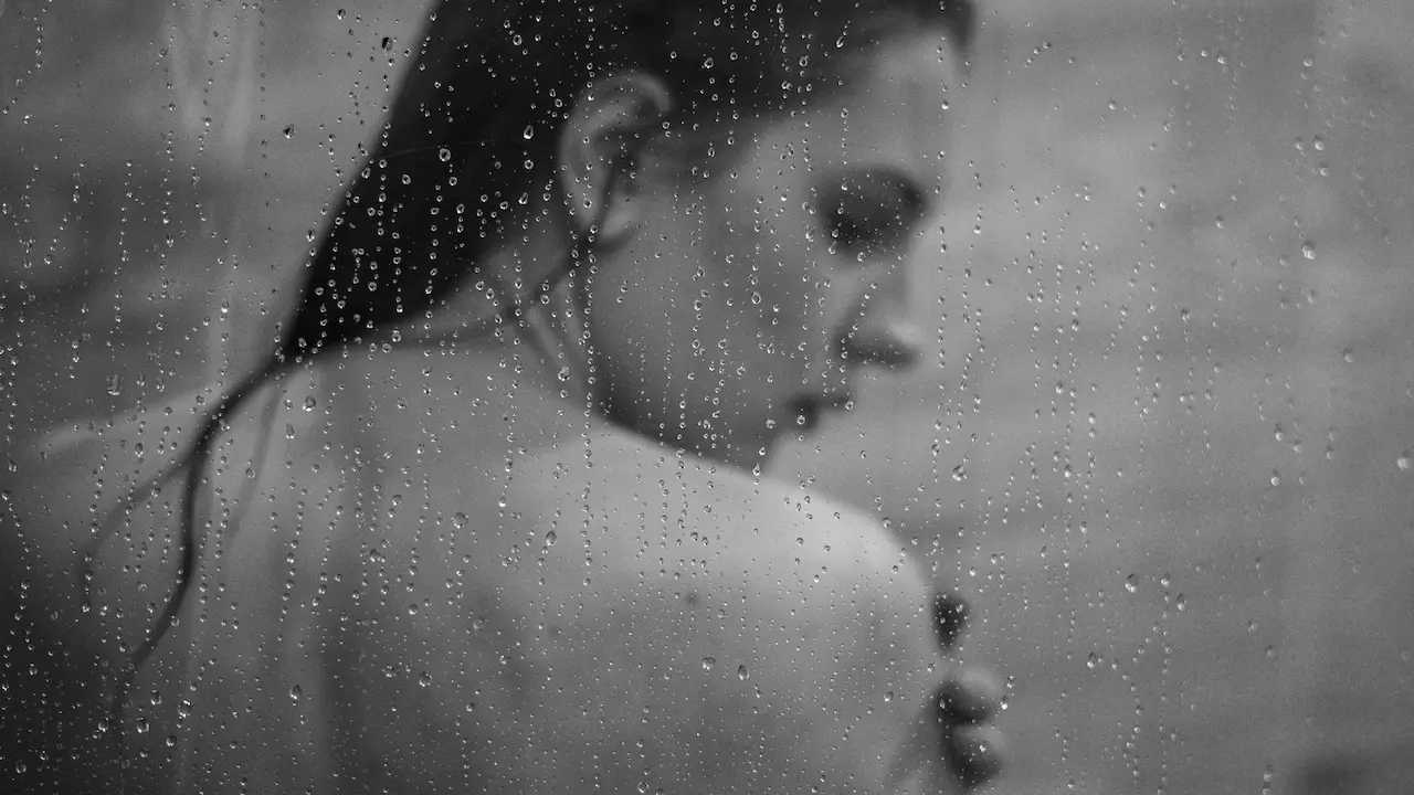 Woman seen through a glass shower screen covered in water droplets, with wet hair, in black and white.